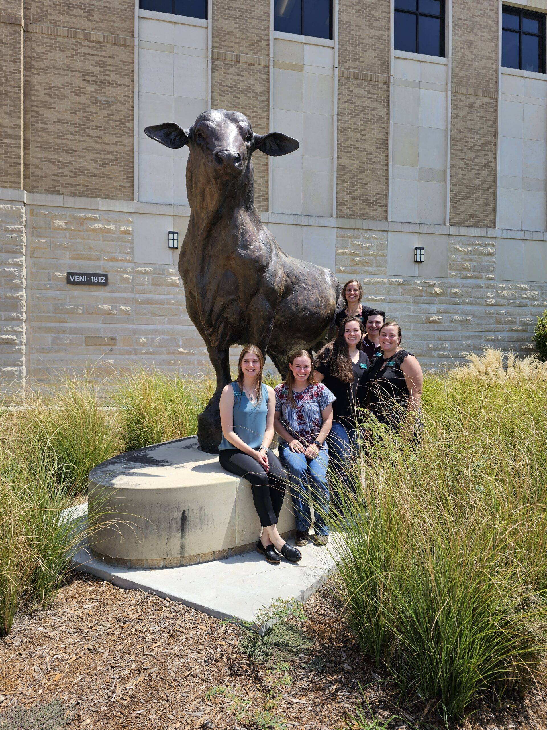 Five women and one man gathered around a large bronze sheep statue outdoors.