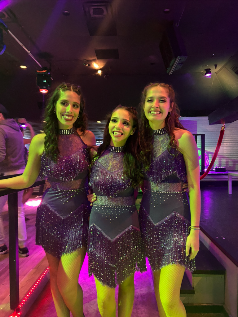 Three young women in purple glittery Latin dance costumes under purple and yellow lights.