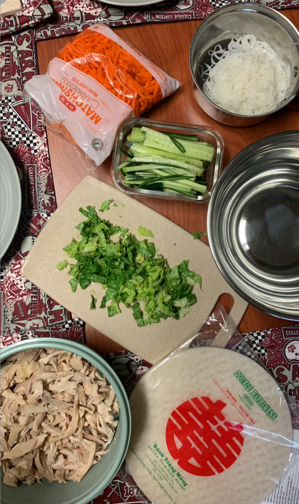 Close up of a table covered in healthy cooking ingredients, including chicken, tortillas, cucumbers, carrots, and chopped greens.