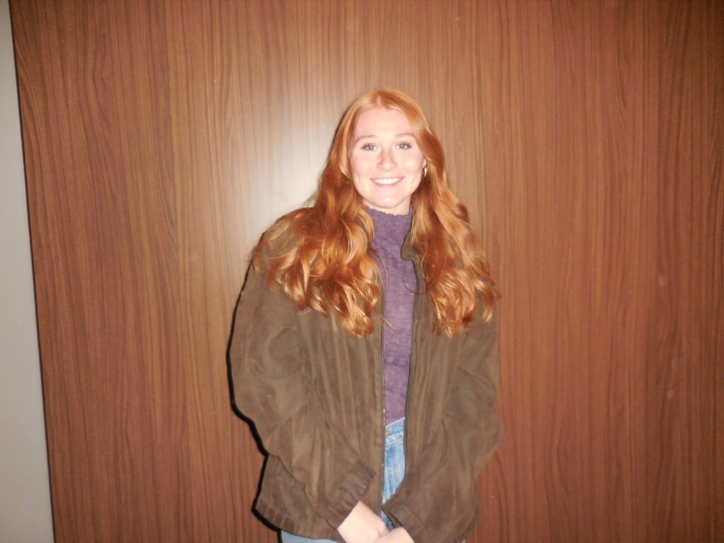 A woman with red hair wearing a brown jacket in front of a wooden wall.