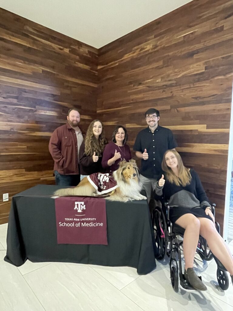 Five individuals, one in a wheelchair, pose for a photo with Reveille, a dog who is the Texas A&M Mascot.