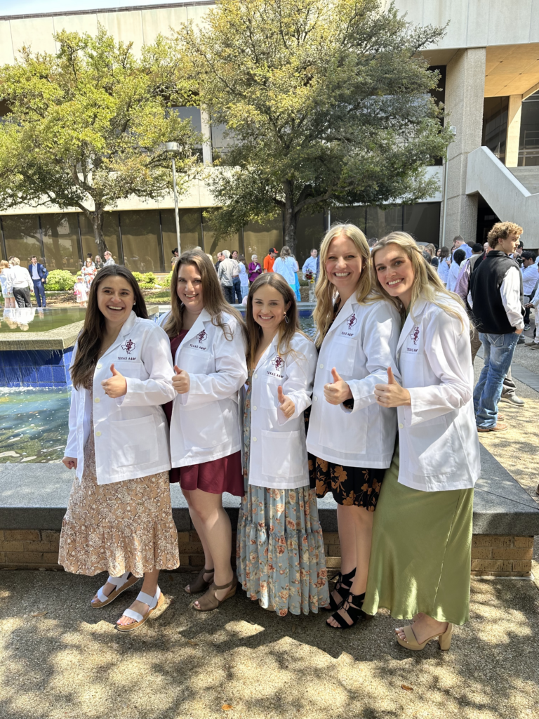 Five young women wearing white physician lab coats standing in a line with their thumbs up.