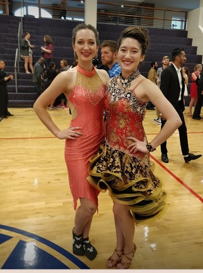 Two young women in ballroom dancing dresses posing for a photo in a school gym.