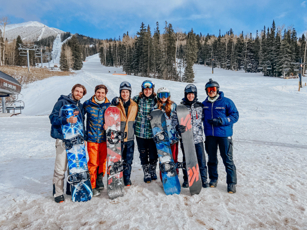 Seven college students in snowboarding gear standing in front of a snowy slope with pine trees.