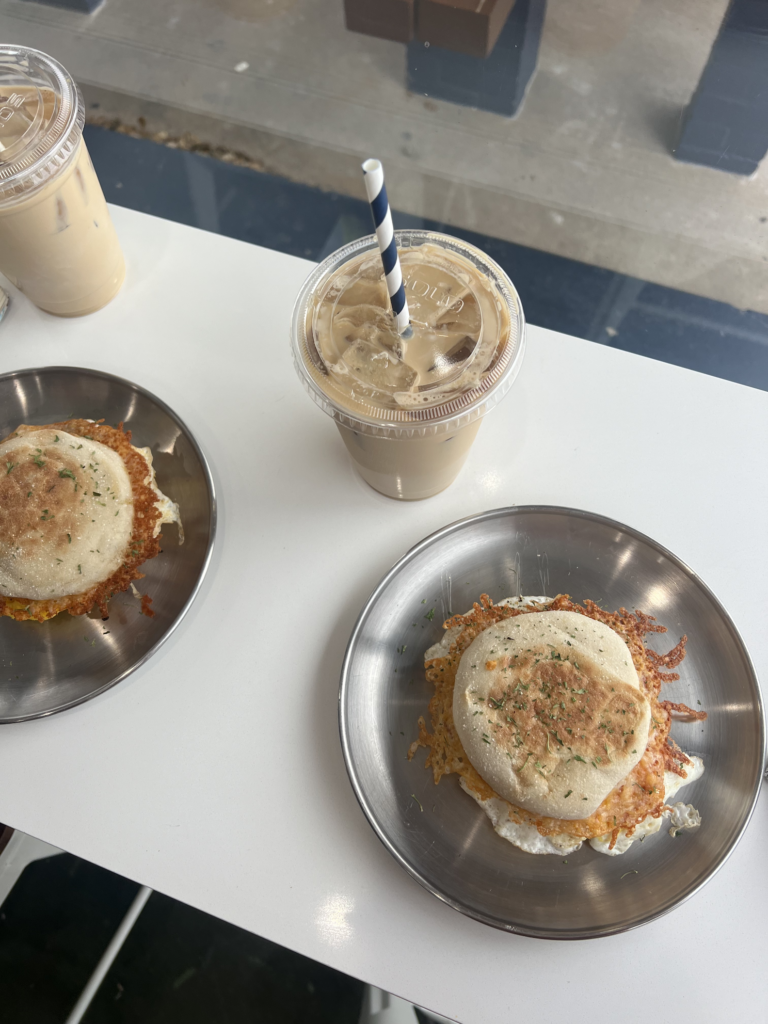 A pair of iced coffee drinks on a white table with two English muffin sandwiches.