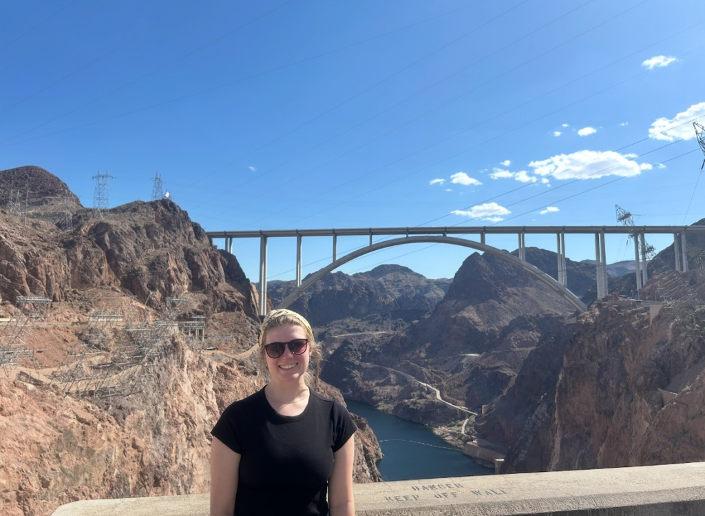 Katie stands smiling, with the Hoover Dam in the background under blue sky