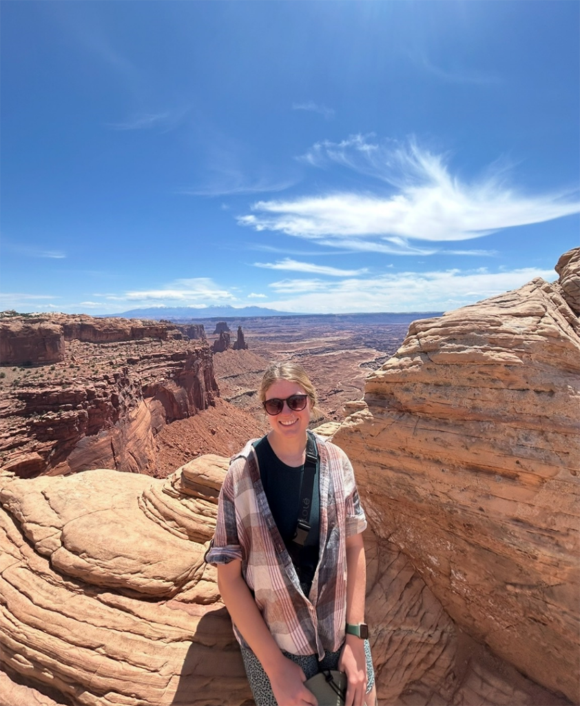 Katie sits, smiling, on a rock with a large canyon in the background under bright blue skies