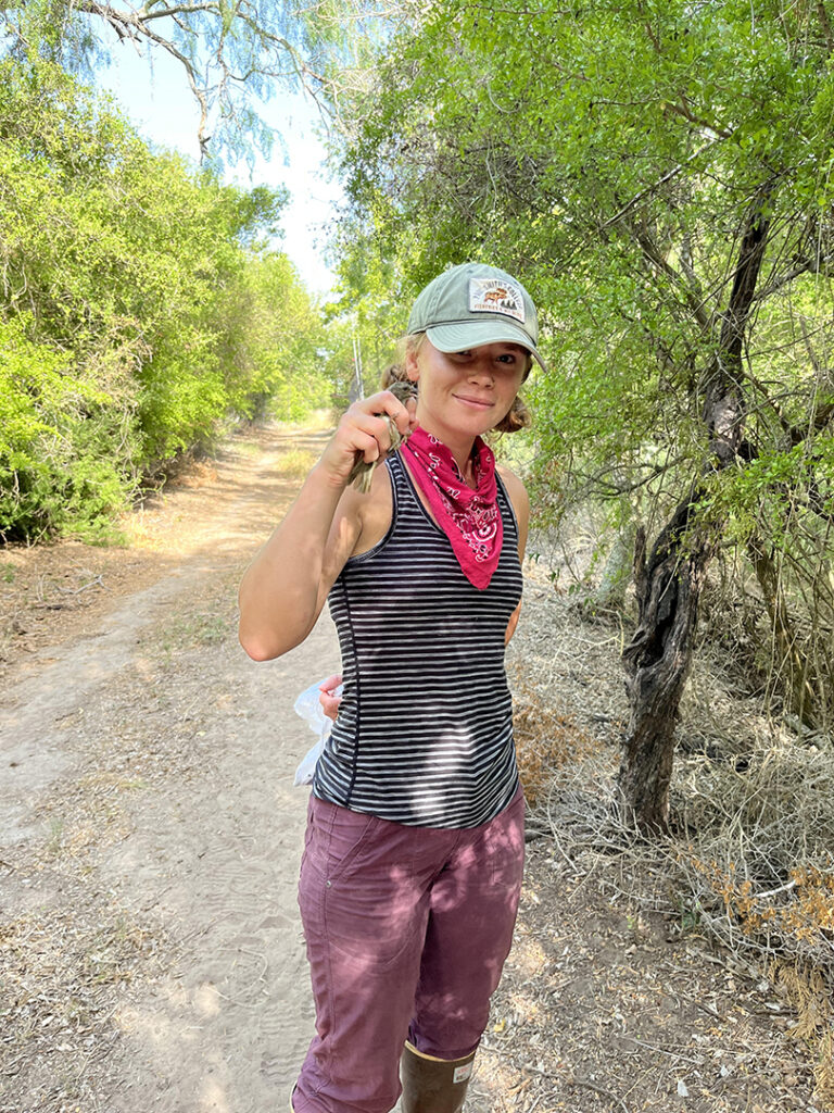 Anna stands holding a bird she banded on a dirt path with trees in the background