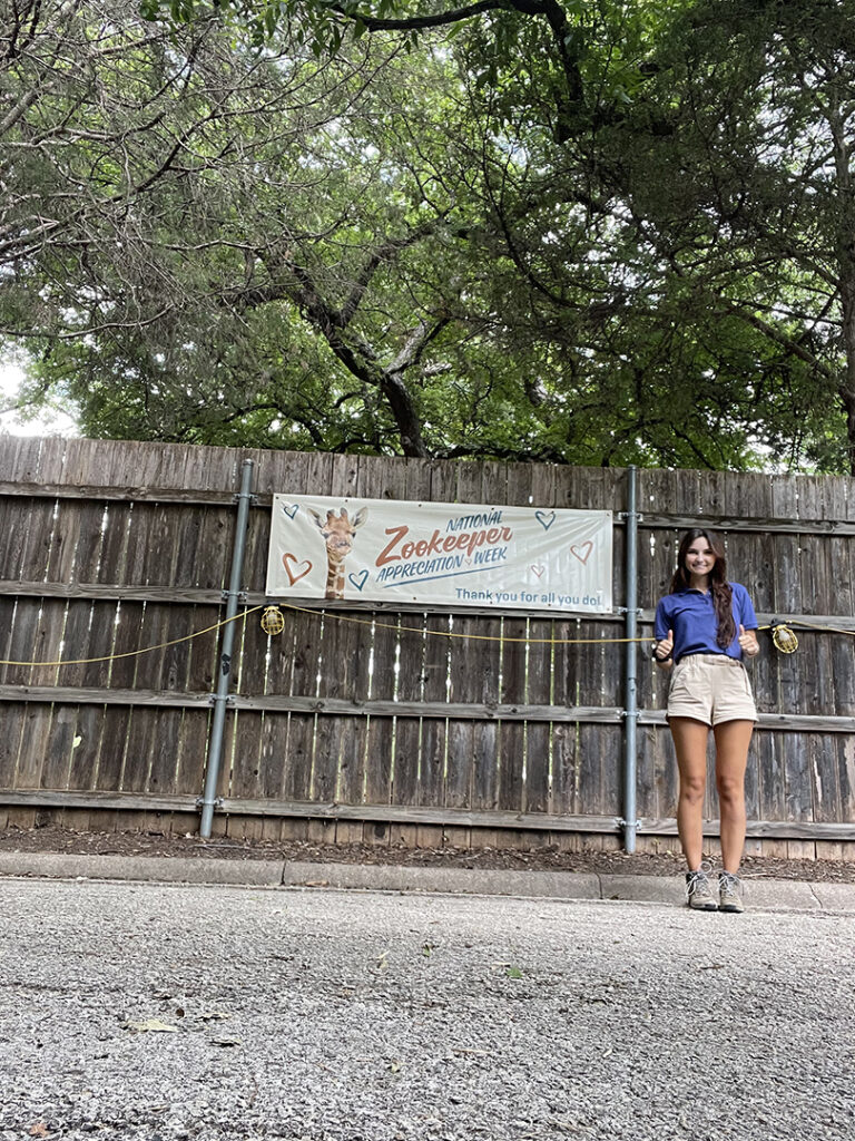 Claire stands with thumbs up in front of a banner that reads "National Zookeeper Appreciation Week" with a graphic of a giraffe, held up on a wood fence.
