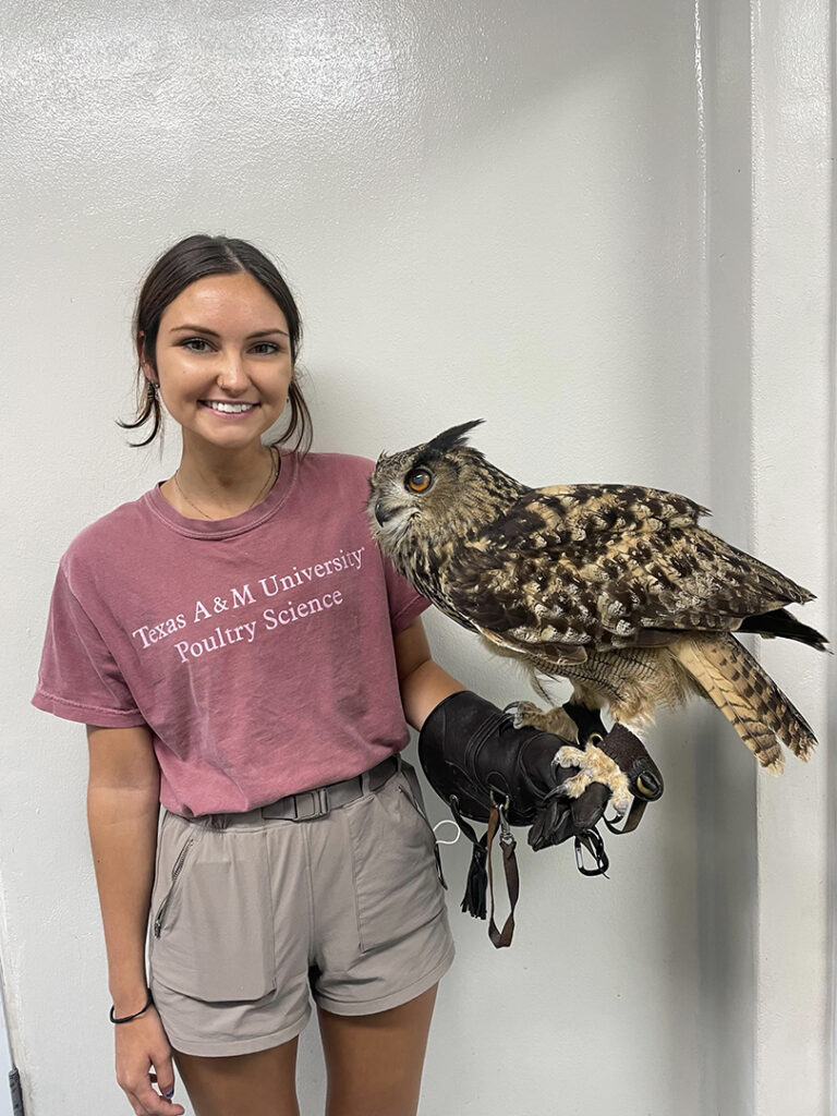 Claire stands, smiling, holding an owl on a gloved hand in front of a gray wall.