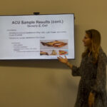 Emma presents her research in a classroom with a screen next to her that displays her research findings.