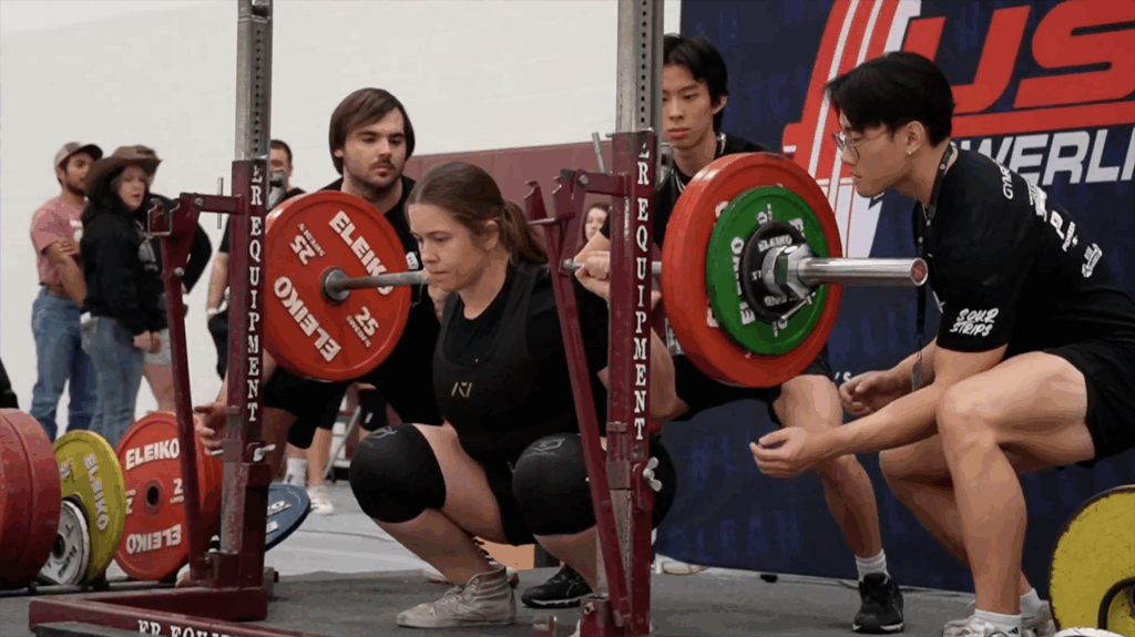 Sarina squats with a barbell and weights in a powerlifting competition with spotters standing by assisting.