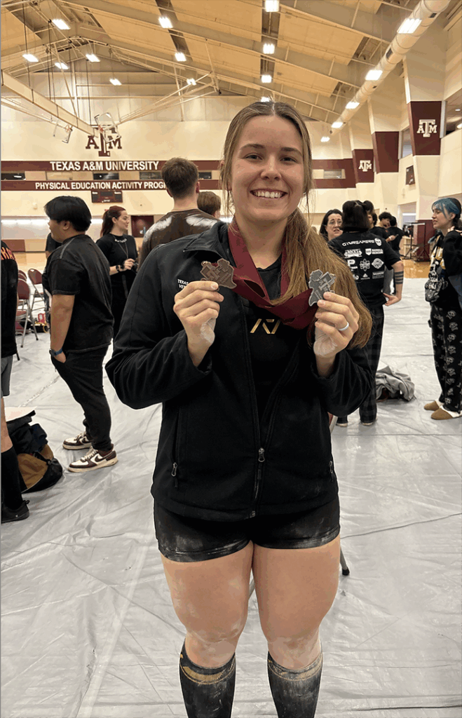 Sarina stands, smiling, while holding medals in each hand. She in inside a gym and wearing athletic shorts and a black jacket.