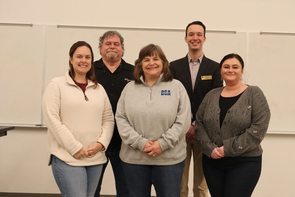 Five adults standing side by side and smiling for a group photo in front of a whiteboard. One person is wearing a badge with the logo "BSA.