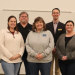 Five adults standing side by side and smiling for a group photo in front of a whiteboard. One person is wearing a badge with the logo "BSA.