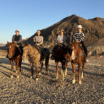 Four individuals riding horses in a desert landscape with mountains in the background.