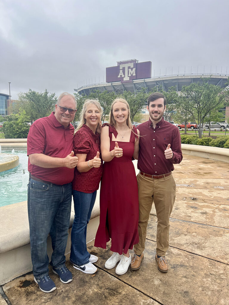 Sarah stands with three individuals, all giving thumbs up, with the Texas A&M University stadium in the background.
