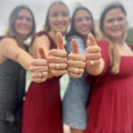 Four people smiling and giving a thumbs up to the camera, showcasing rings on their thumbs, with a focus on the rings and a blurred background. They are dressed in formal attire.