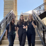 Three veterinary students stand in scrubs and muck boots in front of an exterior staircase