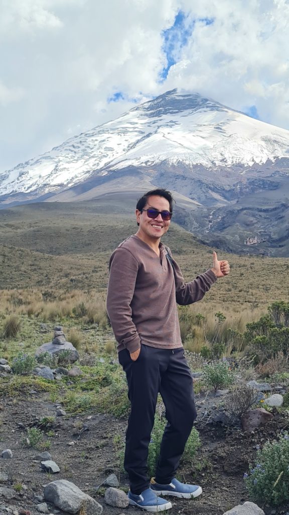 A smiling man with dark hair and sunglasses stands in front of Cotopaxi, a glacier-covered stratovolcano in the Ecuadoran Andes. The man's left hand has his fingers clasped into his palm with his thumb sticking straight up; this gesture is the Texas A&M University sign called "gig 'em."