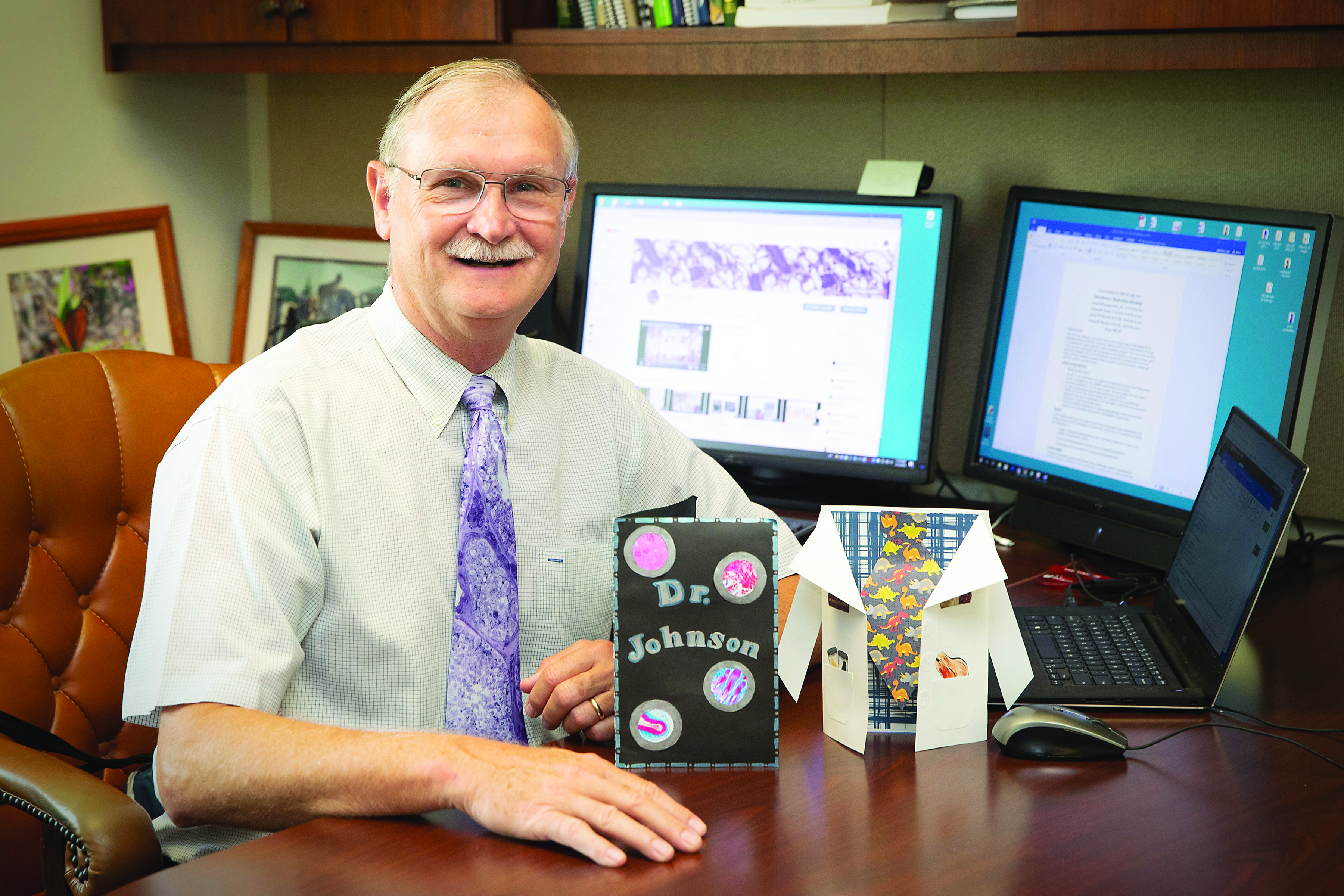 Dr. Larry Johnson, wearing a tie showing a tissue scan, sits at his desk with two handmade thank you cards sitting on the desk in front of him.