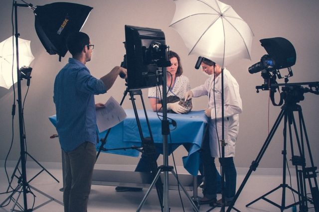 two veterinarians examine a cat on a set while a production crew member adjusts the lighting