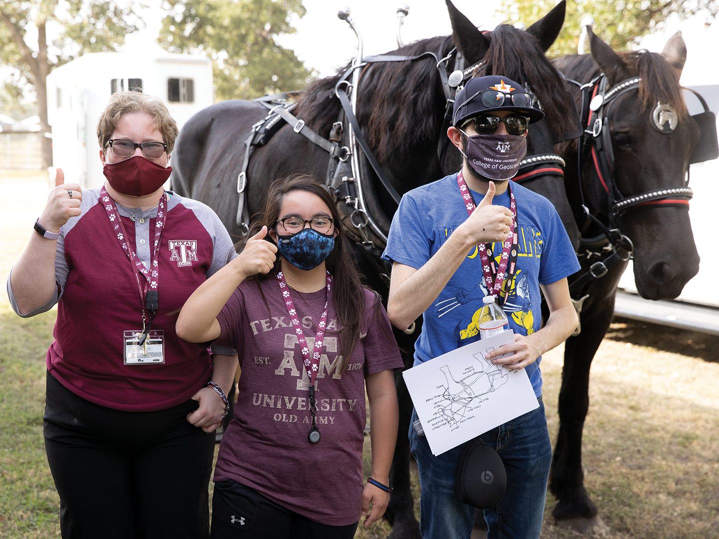 Three students give thumbs up in front of two black draft horses