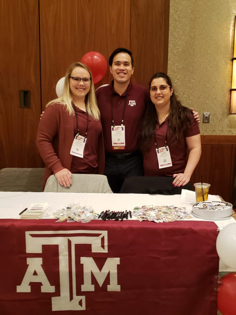 Three people in maroon standing behind a table with a Texas A&M tablecloth