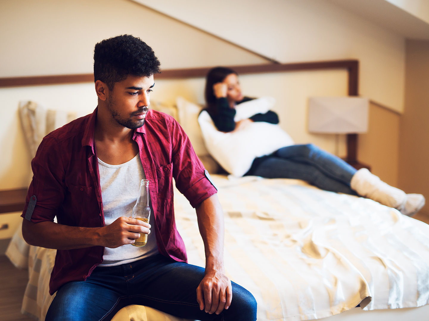 Man drinking alcohol while a sad wife is hugging a pillow in bed