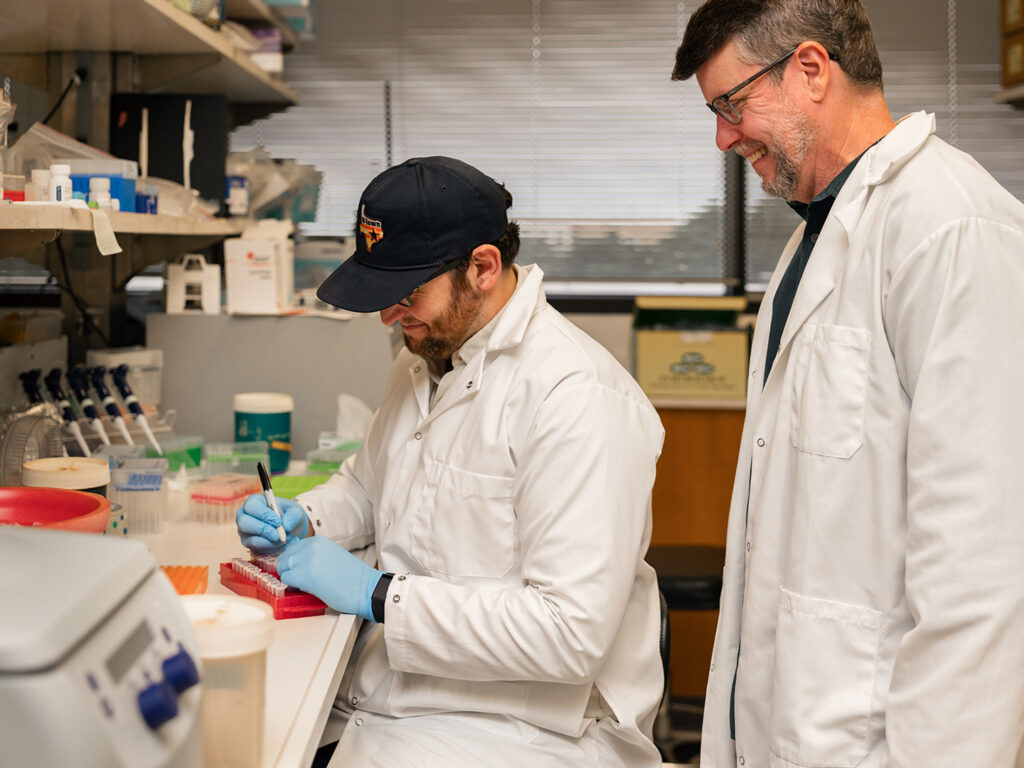 A researcher works with test tubes at a bench while his supervisor watches