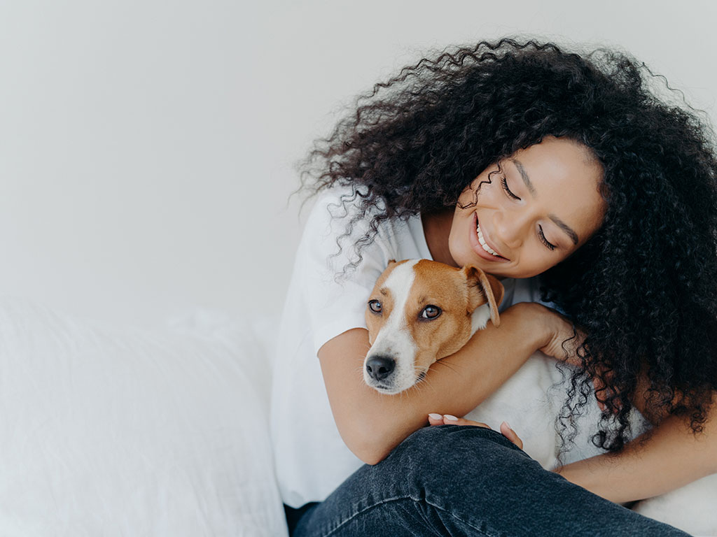 Photo of attractive woman with curly Afro hairstyle, cuddles and pets dog with smile, expresses love, enjoys cozy domestic atmosphere, pose against white background.