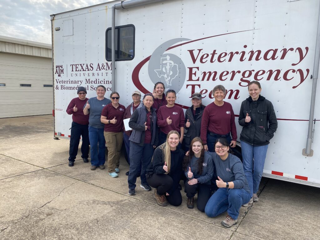 A group of members from the Texas A&M Veterinary Emergency Team stands in front of a disaster response trailer with a white background and a maroon VET logo in maroon. 