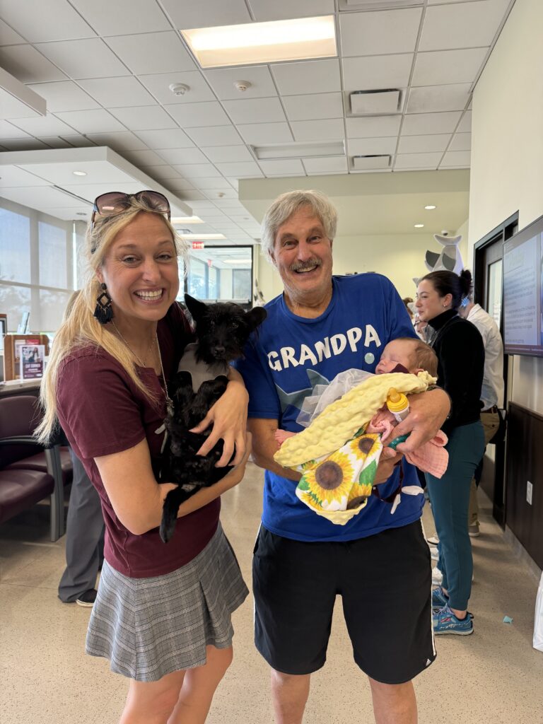 A blonde woman holding a black dog beside a man in a blue shirt holding a newborn baby.