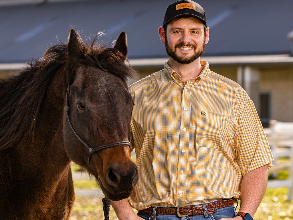 A male veterinary student in a yellow shirt standing beside a brown horse, having won a $75,000 scholarship from Coyote Rock Ranch.