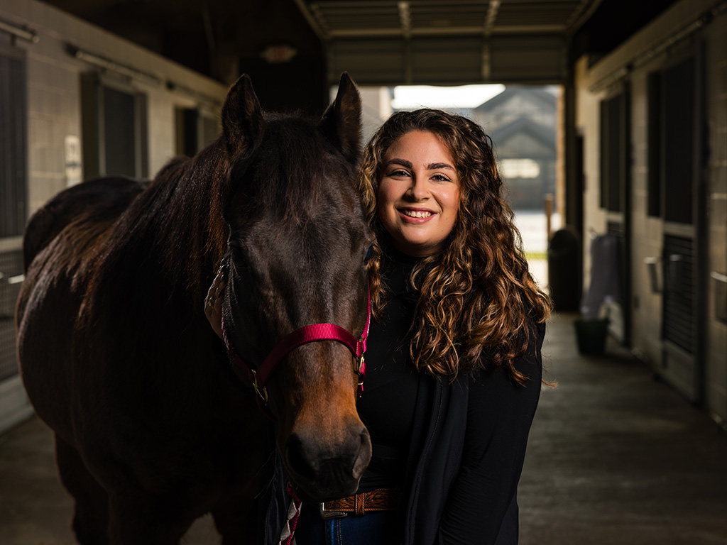 A female veterinary student standing beside a brown horse inside a stable, having won a $75,000 scholarship from Coyote Rock Ranch.