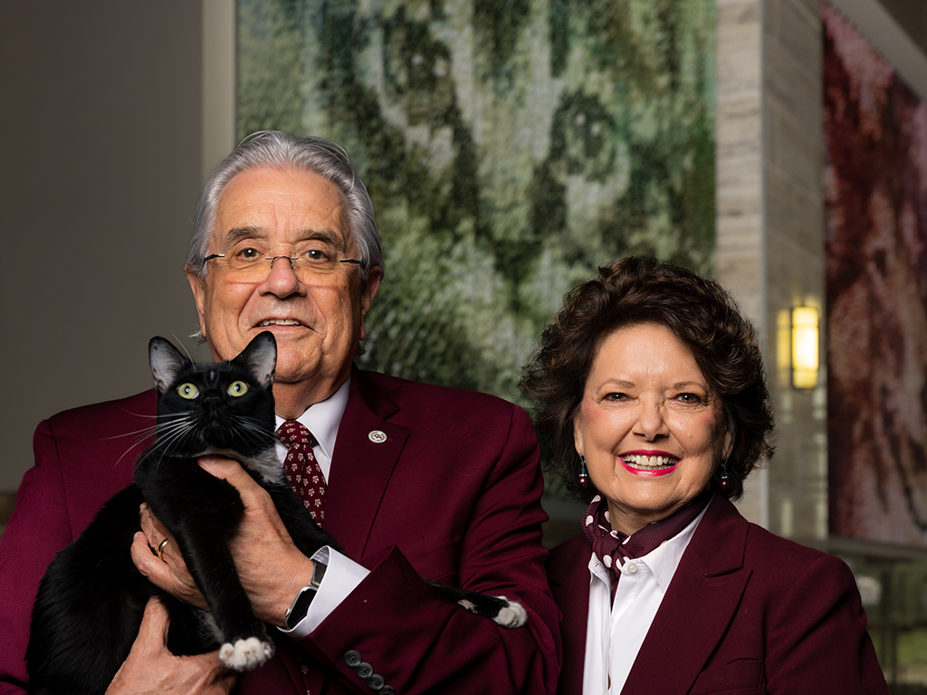A man and a woman in maroon suit jackets and a black cat.